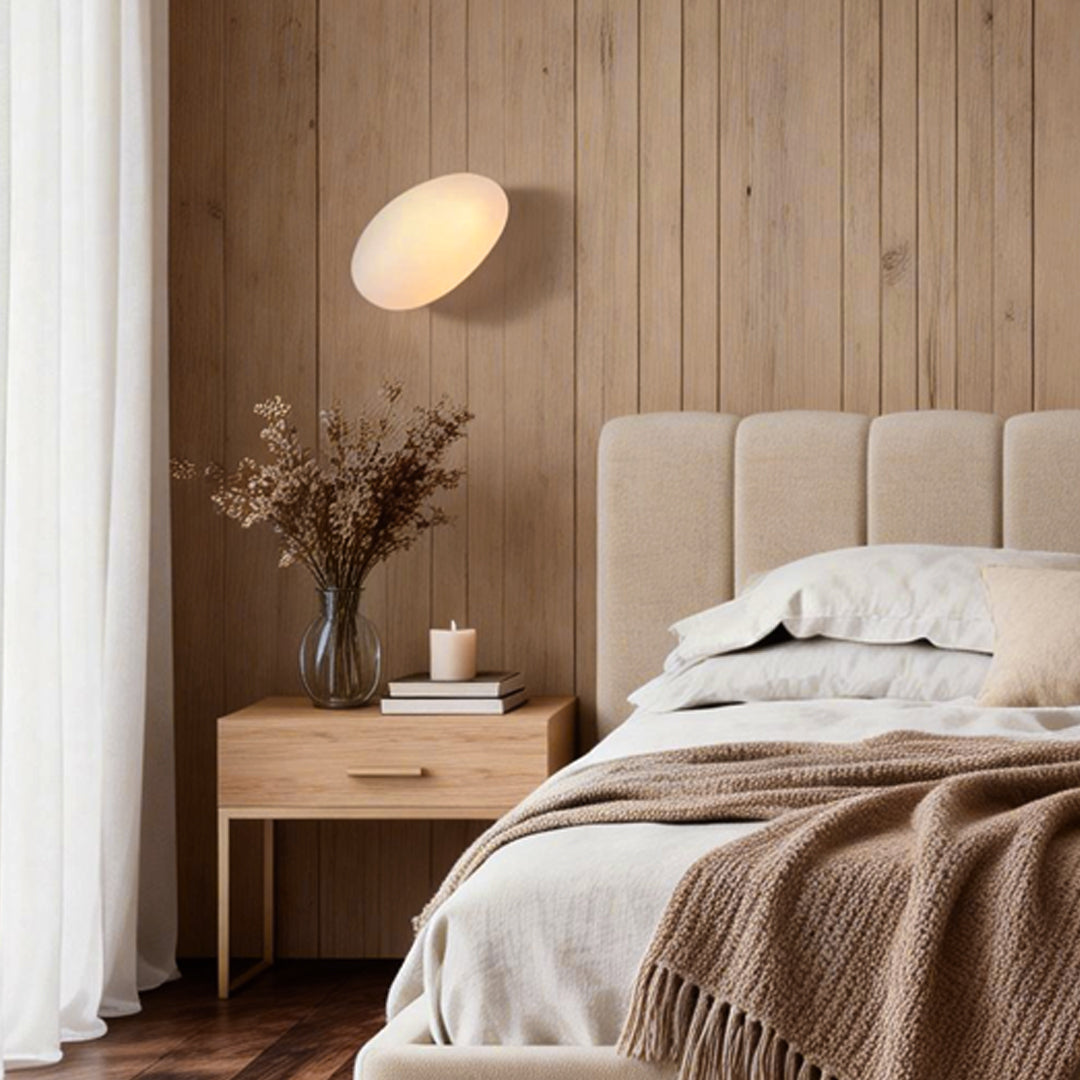 Pebbles wall lamp with a soft glow, mounted above a cozy wooden bed in a modern bedroom.