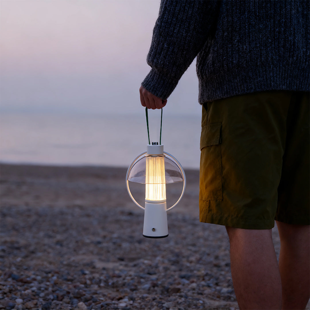 Person holding a white base circle table lamp with a clear shade while standing on a sandy beach at twilight. 