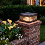 Outdoor pillar mount light placed on a brick column, casting soft light around landscaping and pathway edges at night.