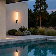 Warm glowing vertical outdoor sconces modern illuminating a white exterior wall by a poolside.