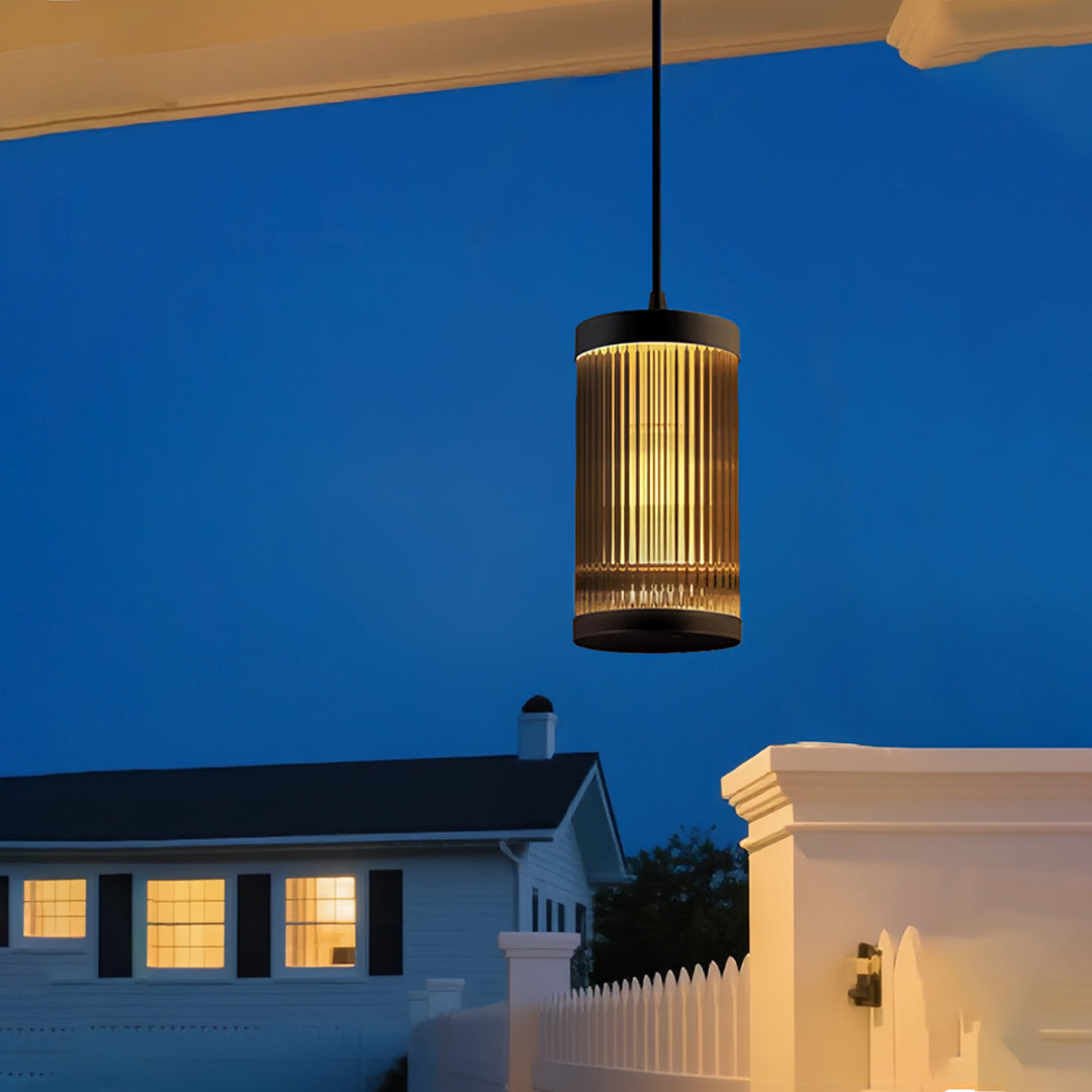 Modern cylindrical outdoor pendant light fixture suspended on a porch at dusk with soft warm illumination.
