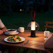 Portable black circle table lamp with a red strap illuminating an outdoor dinner setting on a wooden picnic table. 