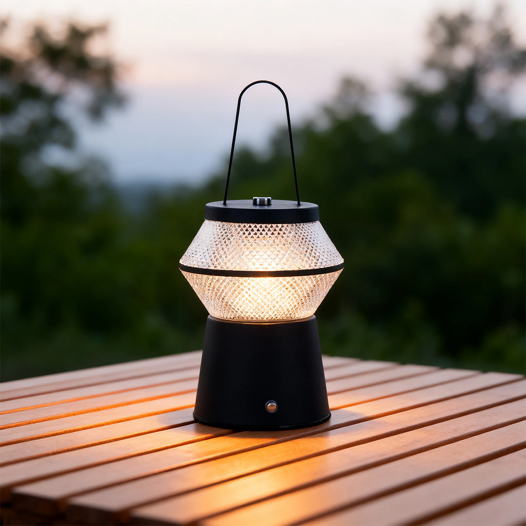 Portable black led outdoor solar lantern sitting on a slatted wooden table outdoors, glowing softly at twilight. 