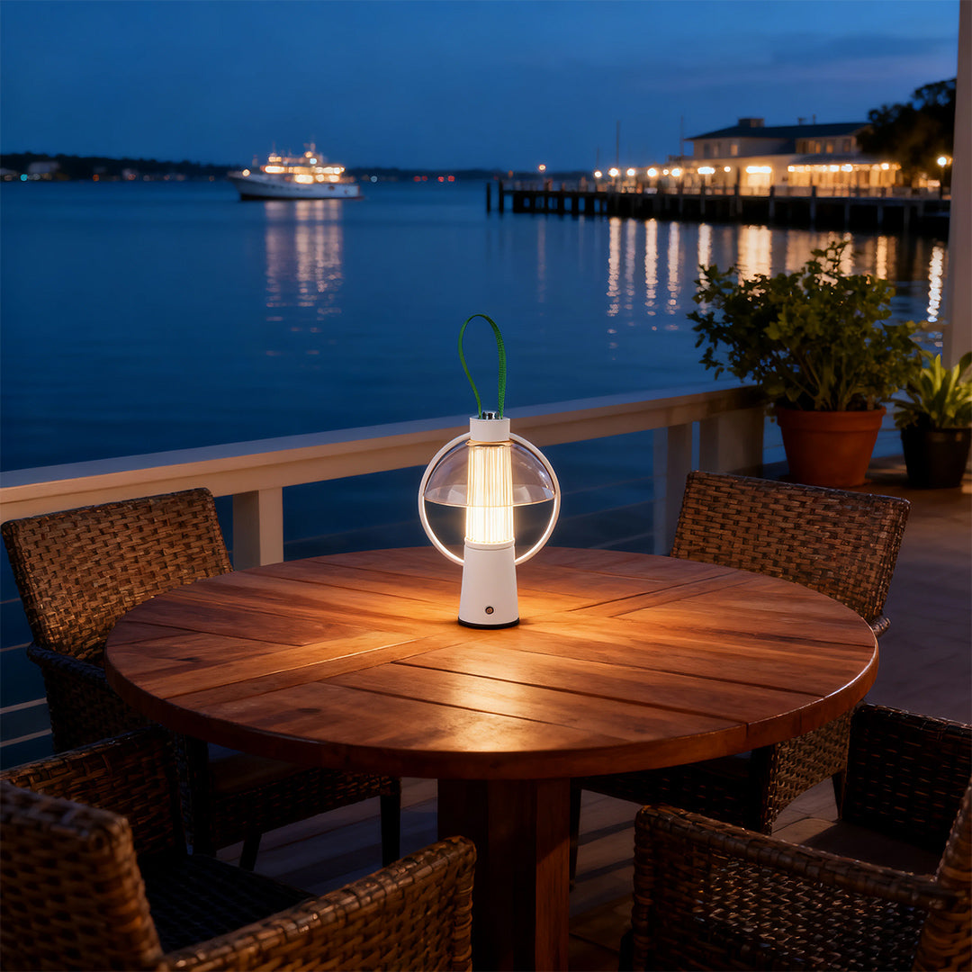 Portable circle table lamp illuminating an outdoor dining table on a balcony overlooking a scenic harbor at night. 