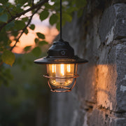 Portable hanging light with a vintage cage design, illuminating a rustic stone wall at dusk.