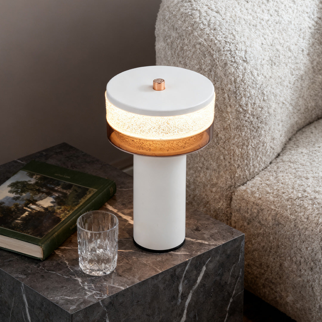 Portable white cylindrical table lamp with a textured shade illuminating a marble side table next to a plush chair.
