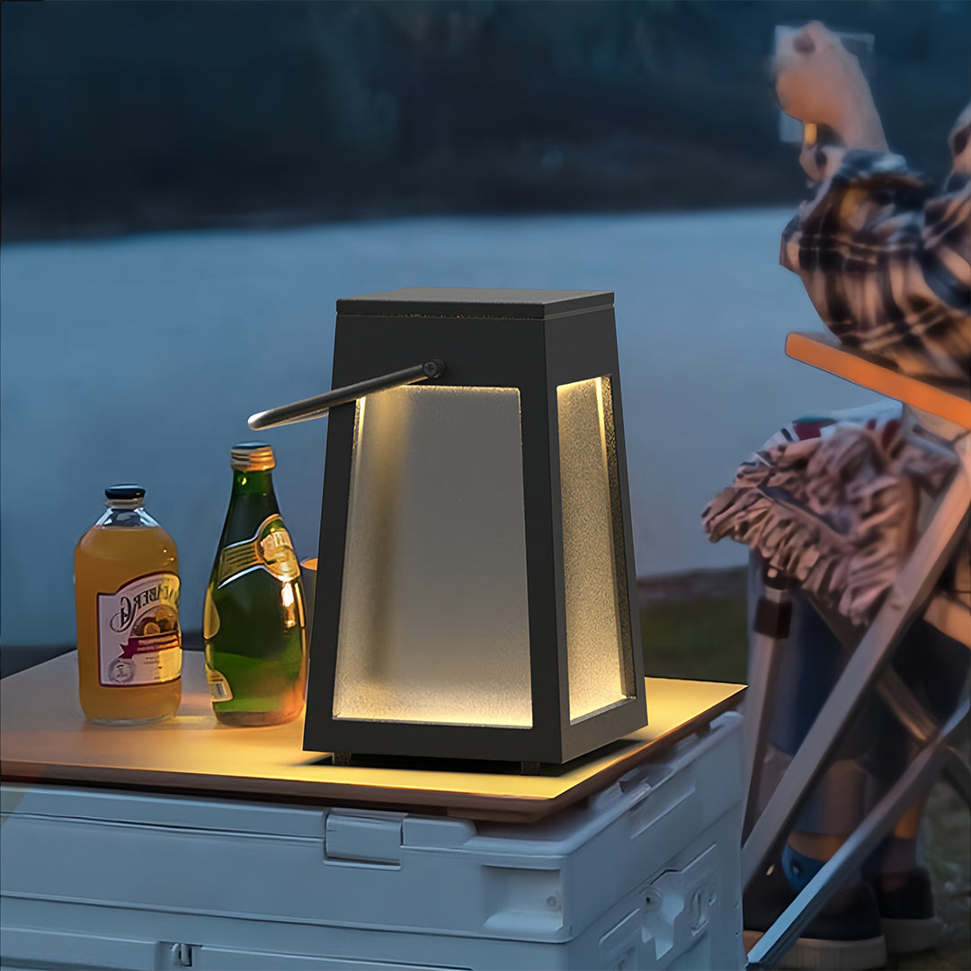Portable camping lantern placed on a wooden picnic table with drinks, used for ambient lighting during an evening outdoor activity.