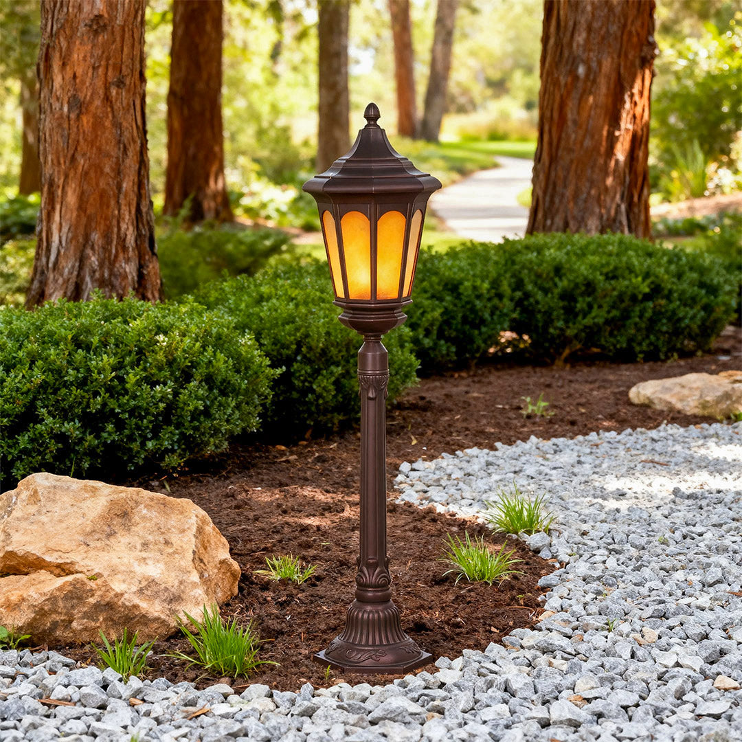 Post path light illuminating garden bed with mulch beside white gravel pathway and trees