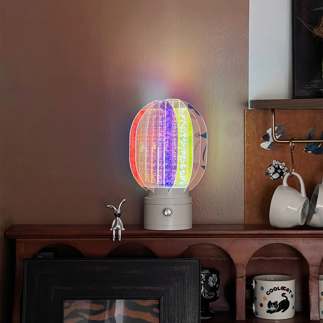 Colourful table lamp casting a rainbow gradient across a cozy bedside area decorated with various artistic paper prints.