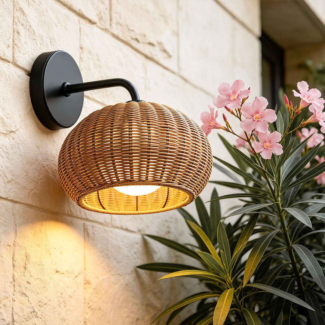 Rattan outdoor pendant light mounted on an exterior wall, glowing warmly beside tropical plants.