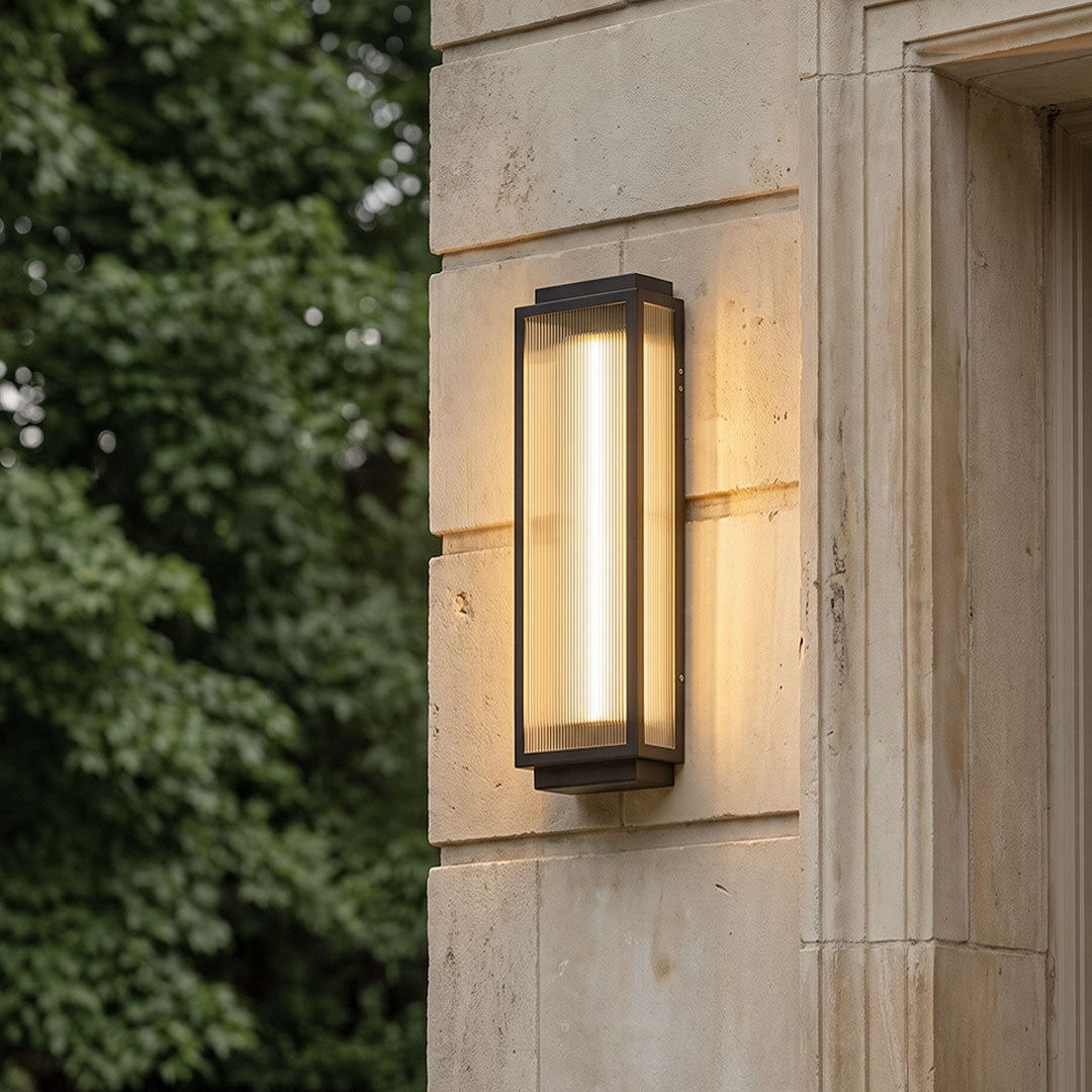 Reeded glass light mounted on a stone exterior wall, featuring a vertical rectangular frame and warm glowing illumination.