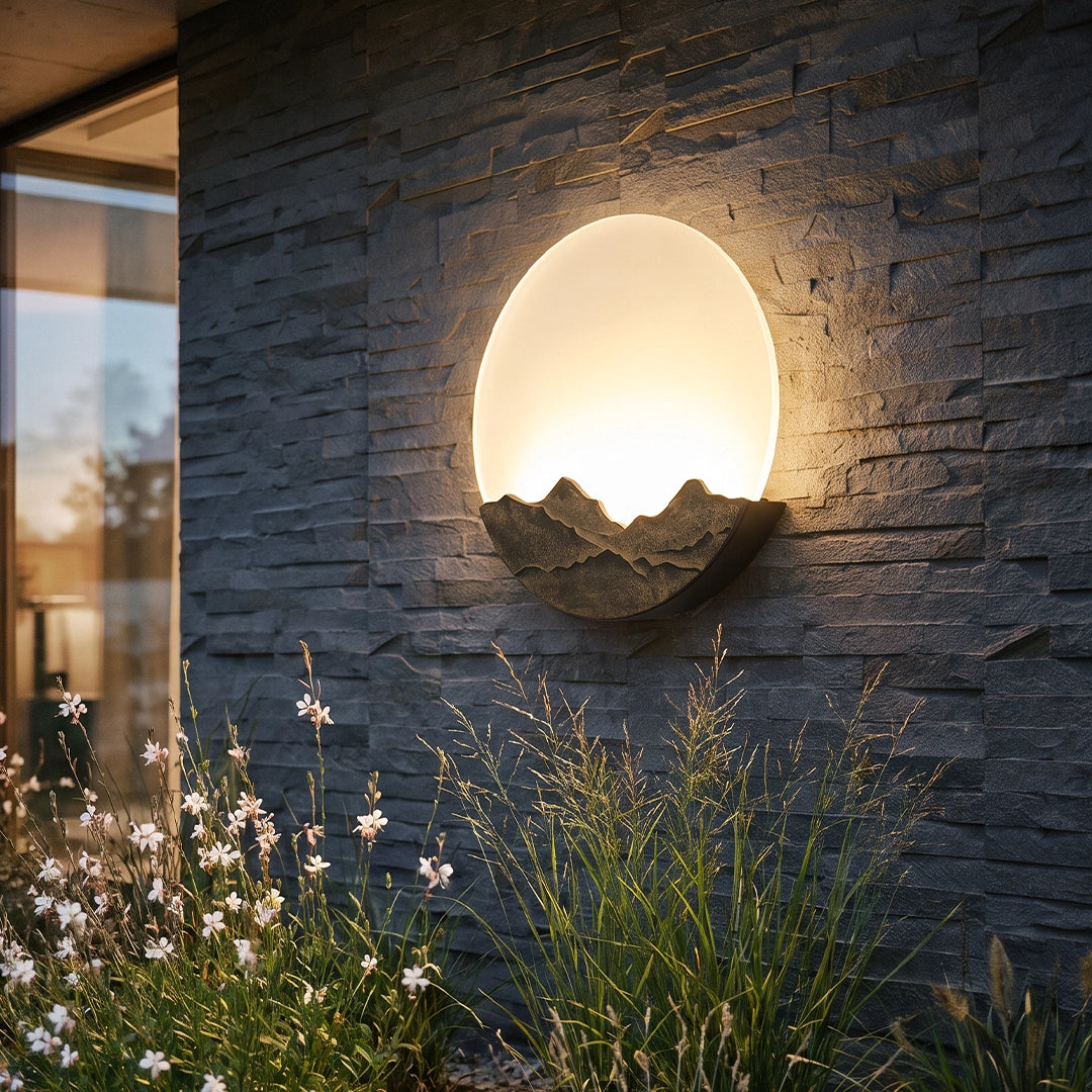 Round outdoor sconce mounted on a stone wall, glowing warmly above garden plants at dusk.