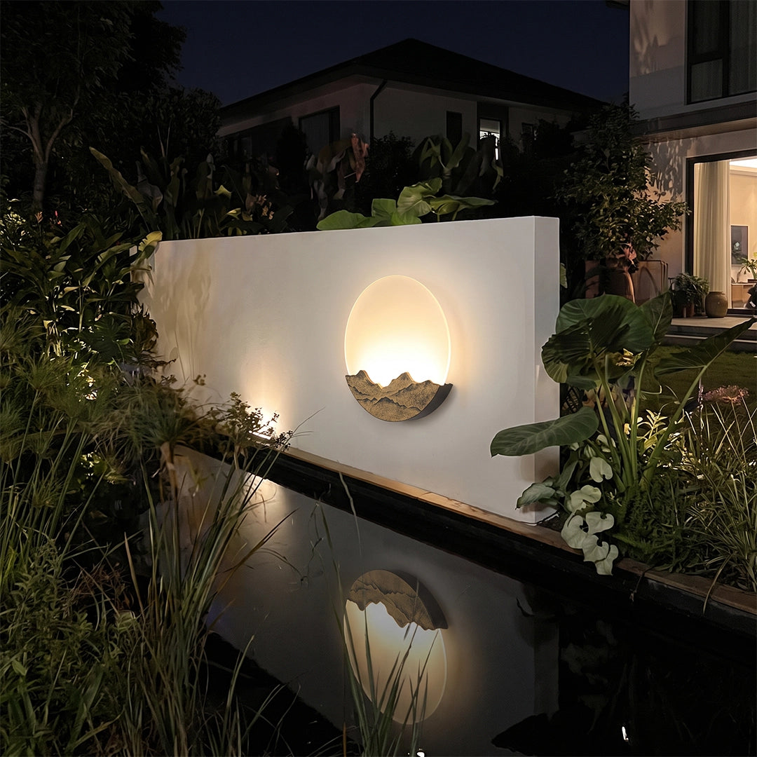 Round outdoor sconce glowing on a white wall beside a reflecting pool and lush plants at night.