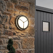 Round outdoor wall light fixture mounted on a rustic stone exterior wall beside a dark entry door.