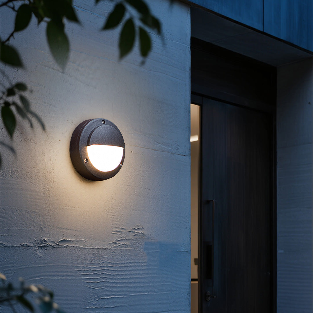 Round outdoor wall sconce illuminating contemporary dark facade at twilight with foliage