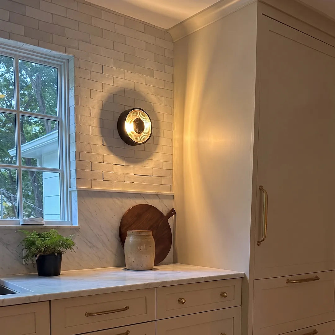 Round wall sconce mounted above a bathroom counter near a window with soft ambient illumination.
