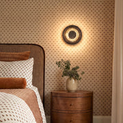 Round wall sconce mounted above a wooden nightstand in a cozy bedroom with patterned wallpaper.