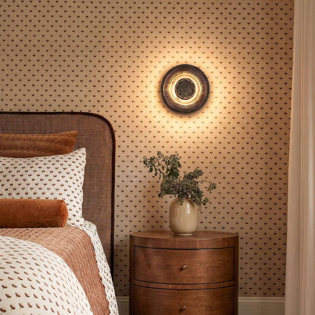 Round wall sconce mounted above a wooden nightstand in a cozy bedroom with patterned wallpaper.
