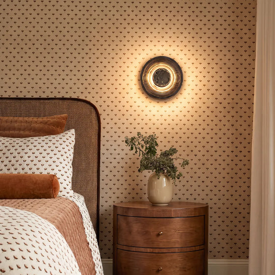 Round wall sconce mounted above a wooden nightstand in a cozy bedroom with patterned wallpaper.