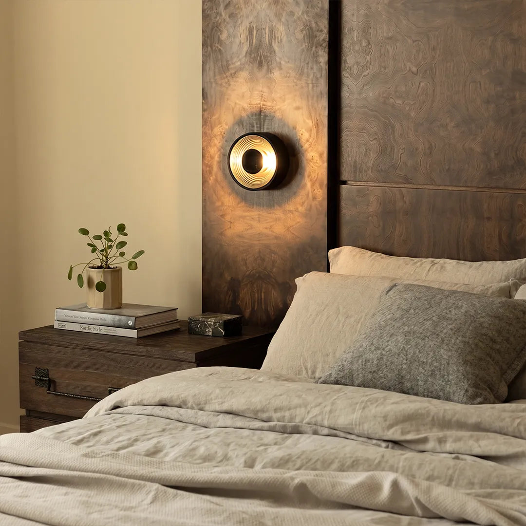 Round wall sconce installed beside a bed with a dark accent wall and warm ambient lighting.