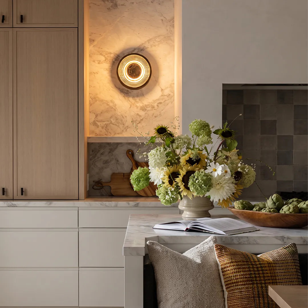 Round wall sconce mounted above a kitchen counter with a textured stone backsplash and warm glow.