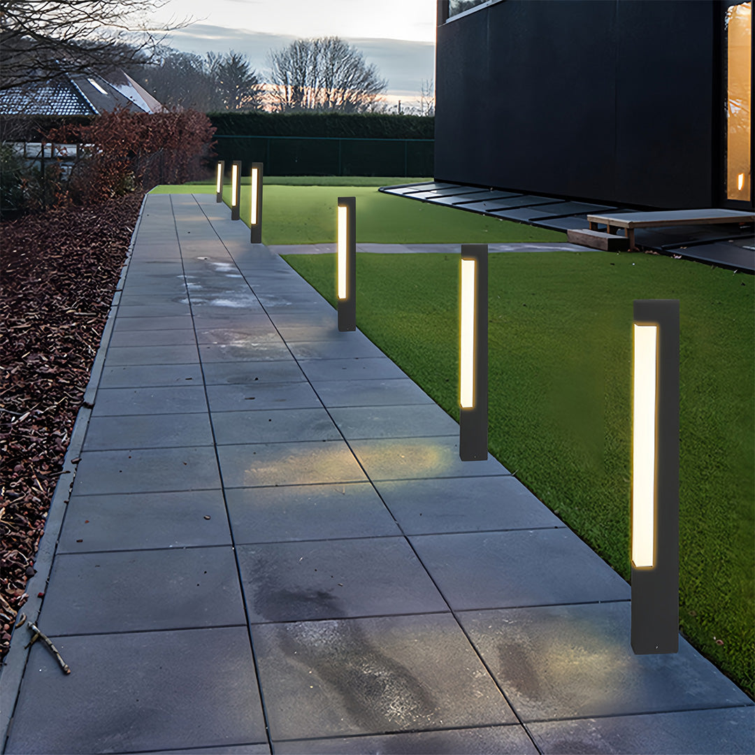 Row of modern black square bollard lights lining a sleek gray stone pathway leading to a contemporary home.