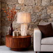 White wood table lamp base with a rustic bark texture, linen shade, placed on a round dark wood side table next to a stone accent wall.