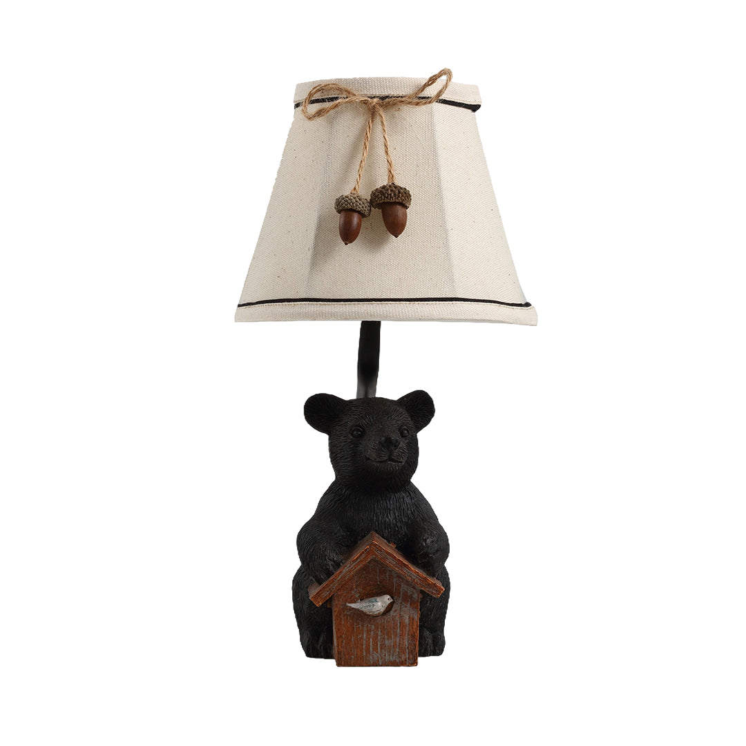 Rustic bear table lamp featuring a carved black bear figurine holding a birdhouse and a neutral fabric shade.