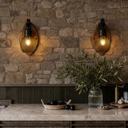Rustic farmhouse wall sconce pair with warm textured glass bulbs and round wood backplates mounted on a stone wall above a kitchen counter.