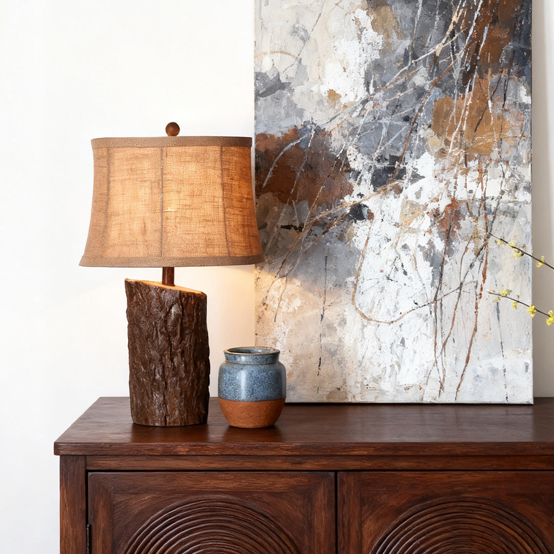 Rustic faux wood table lamp with a split log base and burlap-style linen shade, placed on a dark wood accent cabinet.