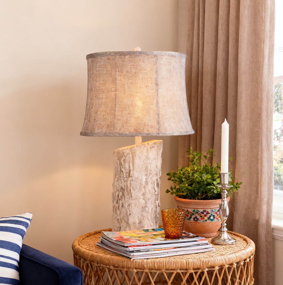 White wood table lamp with a textured tree bark base and a dark, woven fabric shade on a wicker side table next to a window.