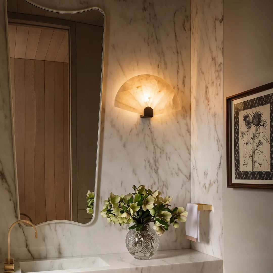 Scalloped wall sconce mounted beside a bathroom mirror with a marble wall and warm ambient lighting.