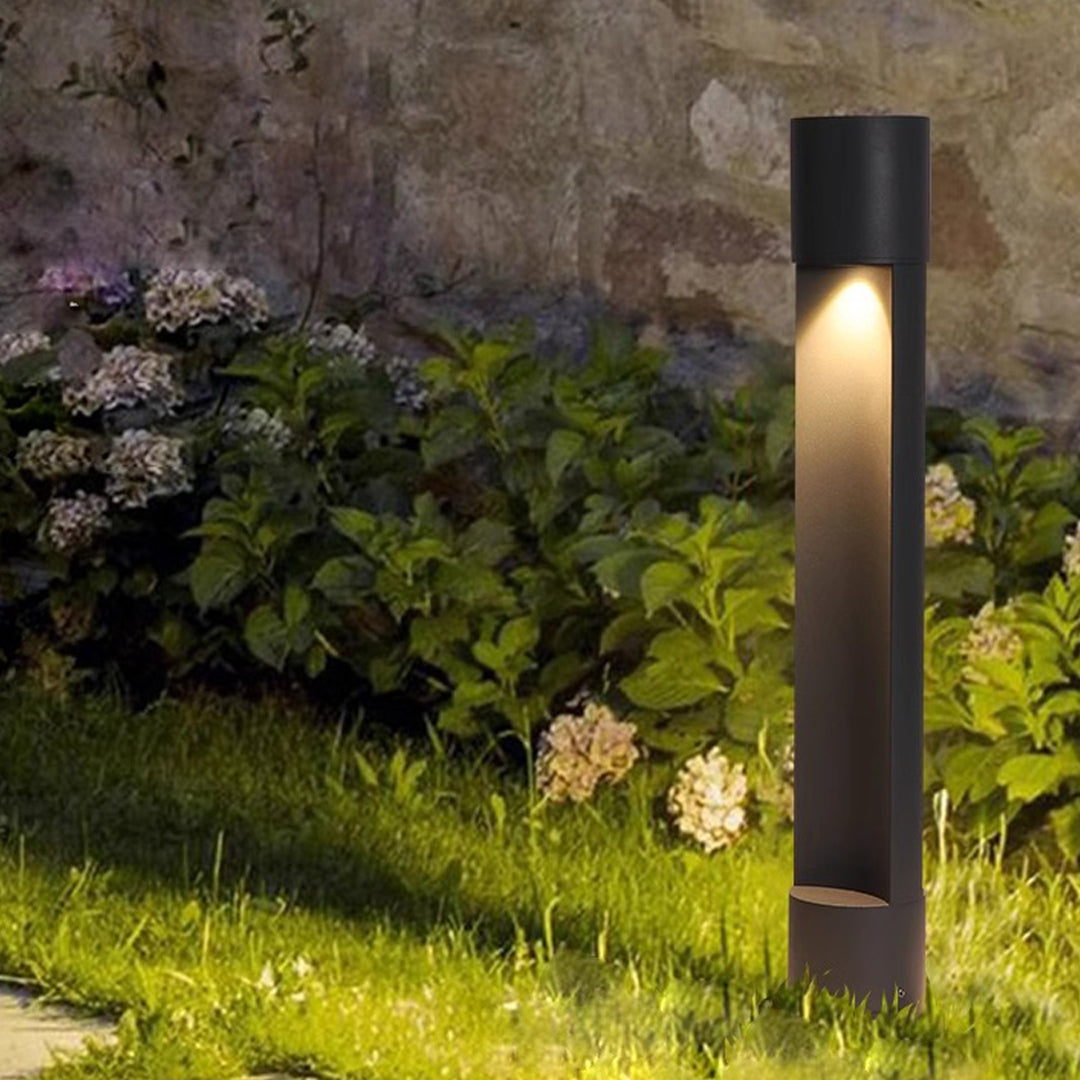 Tall slim sconce outdoor lighting fixture installed in landscaped greenery with soft warm glow.