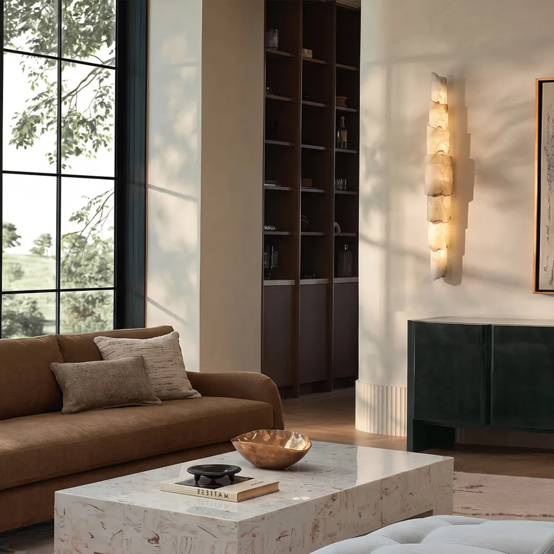 Sculptural wall sconce mounted in a sunlit living room with stacked stone-like shades and a soft warm glow.