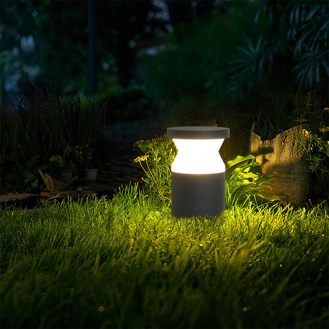Short cylindrical modern path lights illuminating a grassy patch and rock border with a soft, warm LED glow.