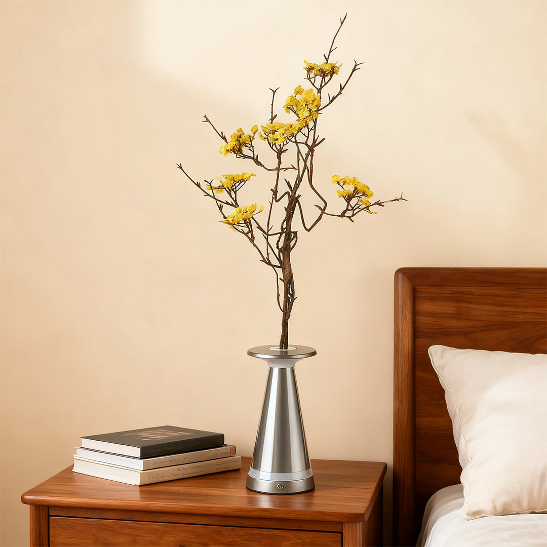 Silver vase table lamp holding yellow flowers on a wooden nightstand in a bright bedroom.