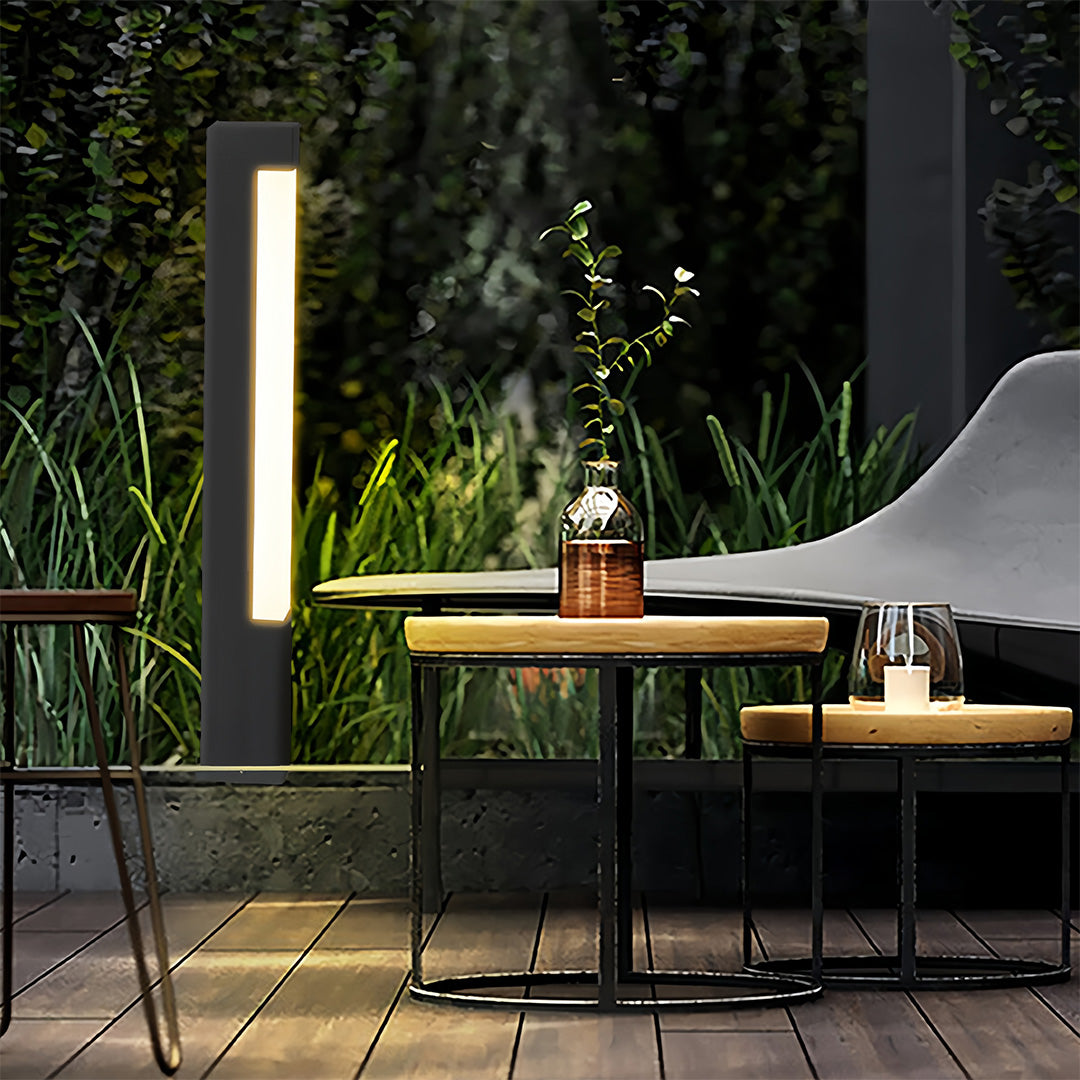 Single square bollard lights fixture next to a wooden outdoor seating area, offering ambient light for a patio corner.