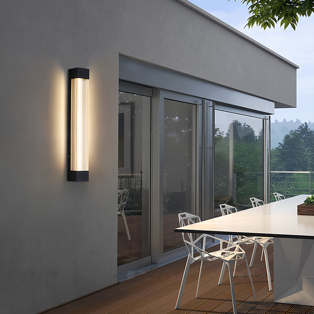 Sleek black linear outdoor sconce providing warm light on a modern patio wall next to a glass door.