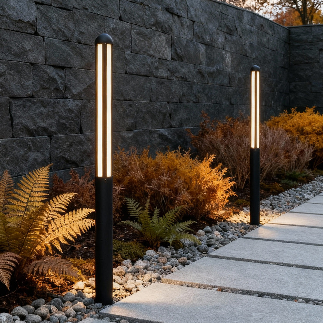 Tall garden bollard light with sleek black design beside stone wall and plants.