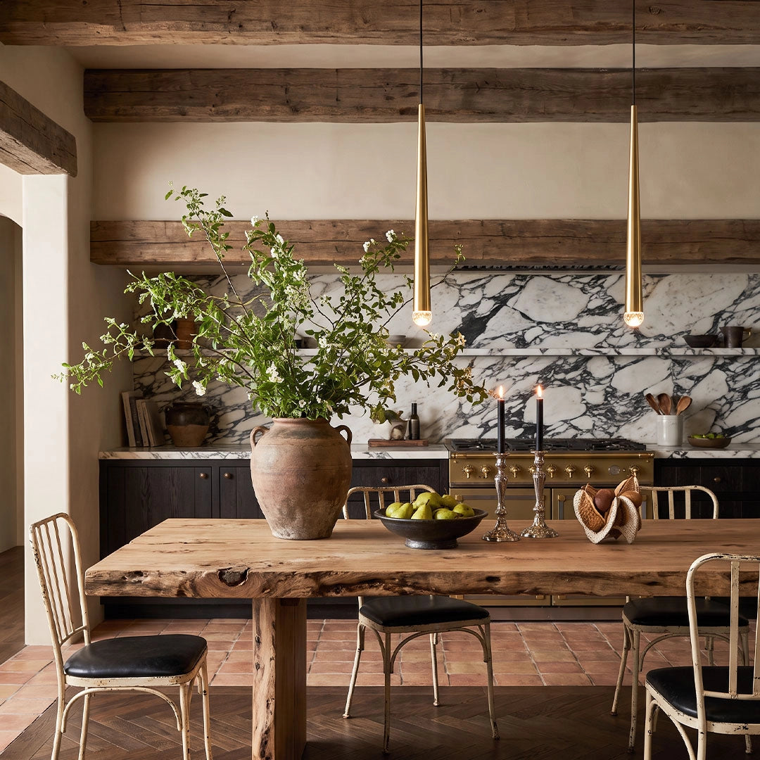 Slim gold cone pendant lights hanging above a rustic dining table in a warm kitchen with exposed wood beams.