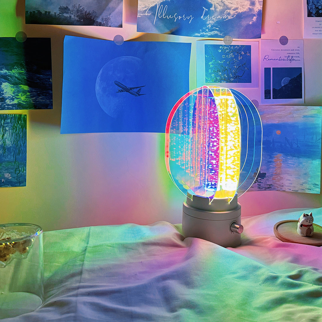 Small oval colourful table lamp sitting on a wooden shelf, providing mood lighting with vibrant projected hues against a dark wall.