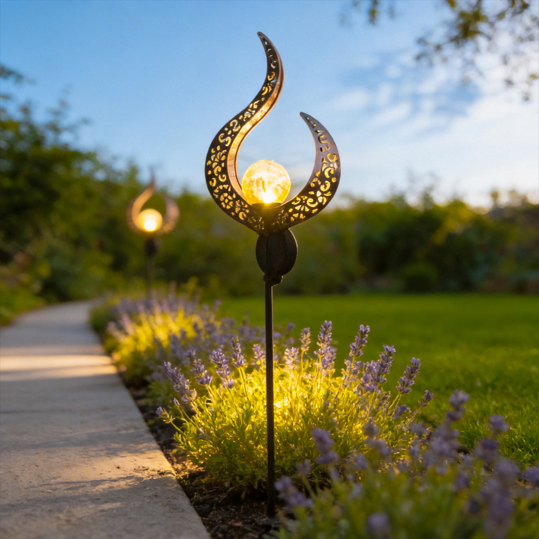Solar garden stake light with a flame-shaped metal shade illuminating flowers beside an outdoor path.