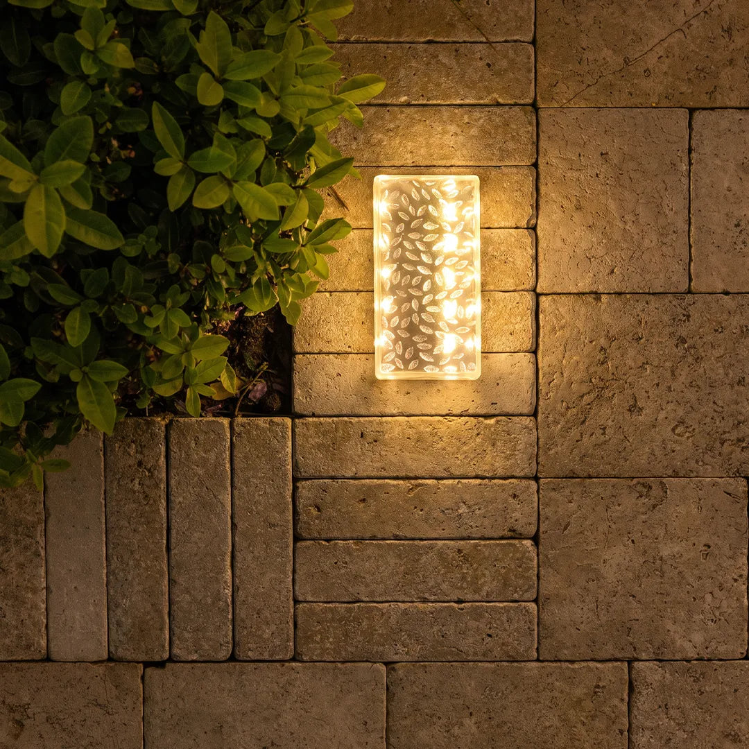 Solar LED step light mounted on a stone wall, glowing beside green shrubs at night.