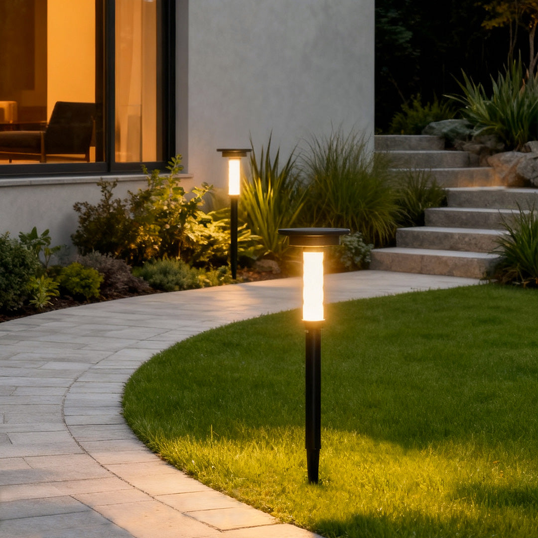 Single solar outdoor path lighting fixture glowing beside grass along a curved residential pathway.