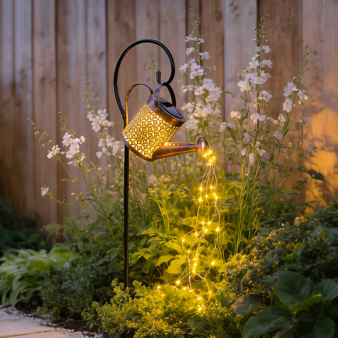 Solar watering can lights cascading warm fairy lights over flowering plants beside a wooden garden fence.