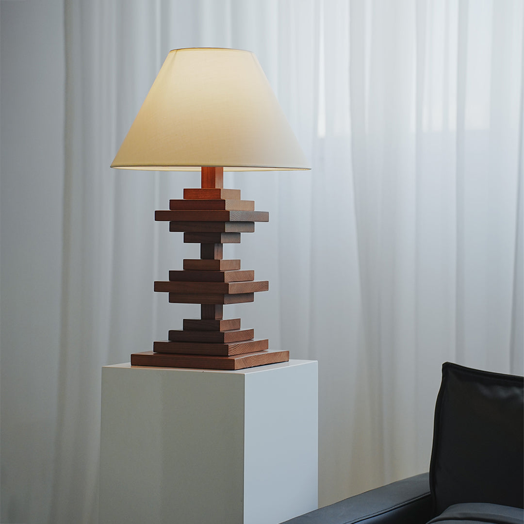 Solid wood table lamps with stacked base placed on white pedestal near curtain.