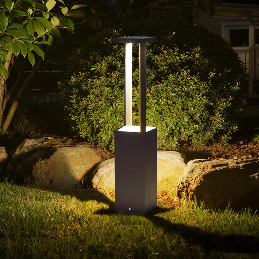 Modern square bollard light fixture positioned among landscaped greenery and natural stones