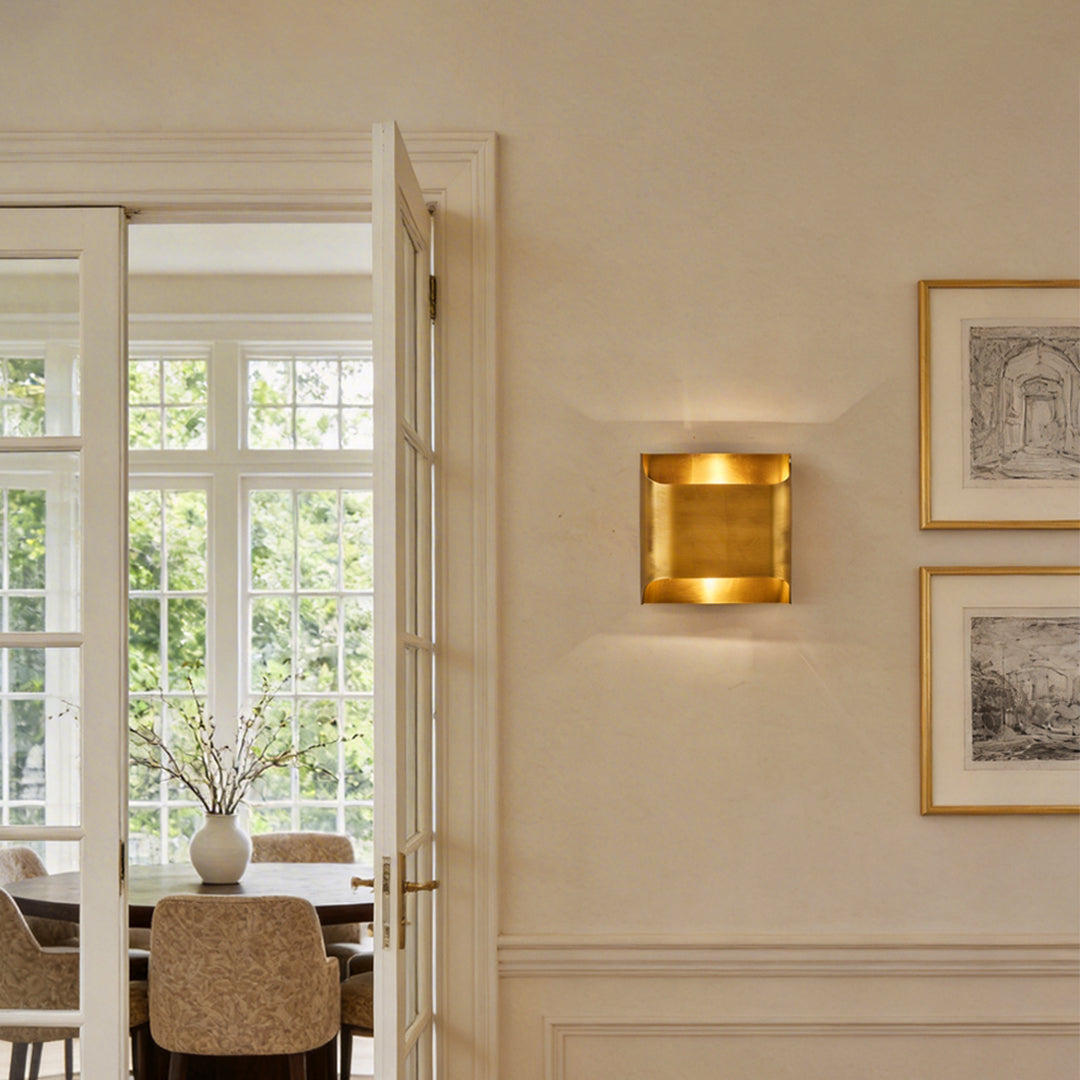 Close up of a square brass wall light glowing warmly on a beige wall in a luxury home interior.