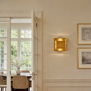 Close up of a square brass wall light glowing warmly on a beige wall in a luxury home interior.