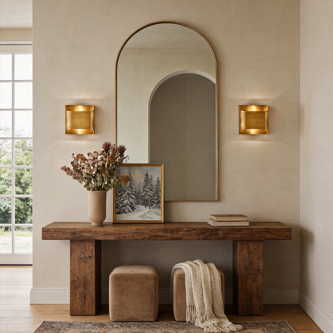 Square brass wall lights illuminating a rustic wood console table with a large arched mirror.